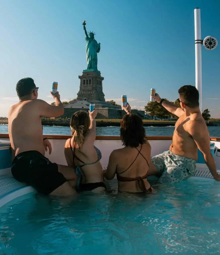 men and women in hot tub boat in front of statue of liberty