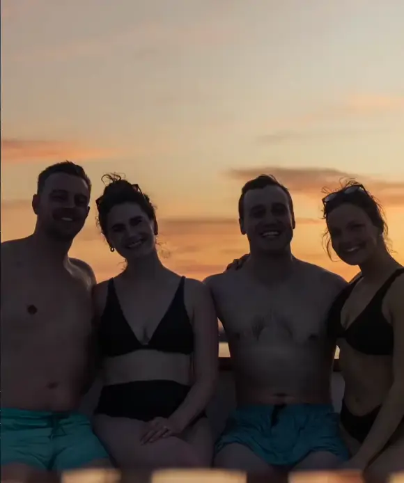 Group of four people in swimsuits posing together on a boat during sunset, with an orange sky over the water and smiles on their faces.